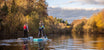 Two women paddleboarding down a river in autumn, the trees either side are orange and then sun is low