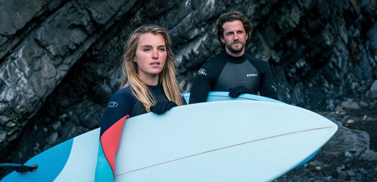 A man and a woman at the beach in winter, holding surfboards and looking past the camera