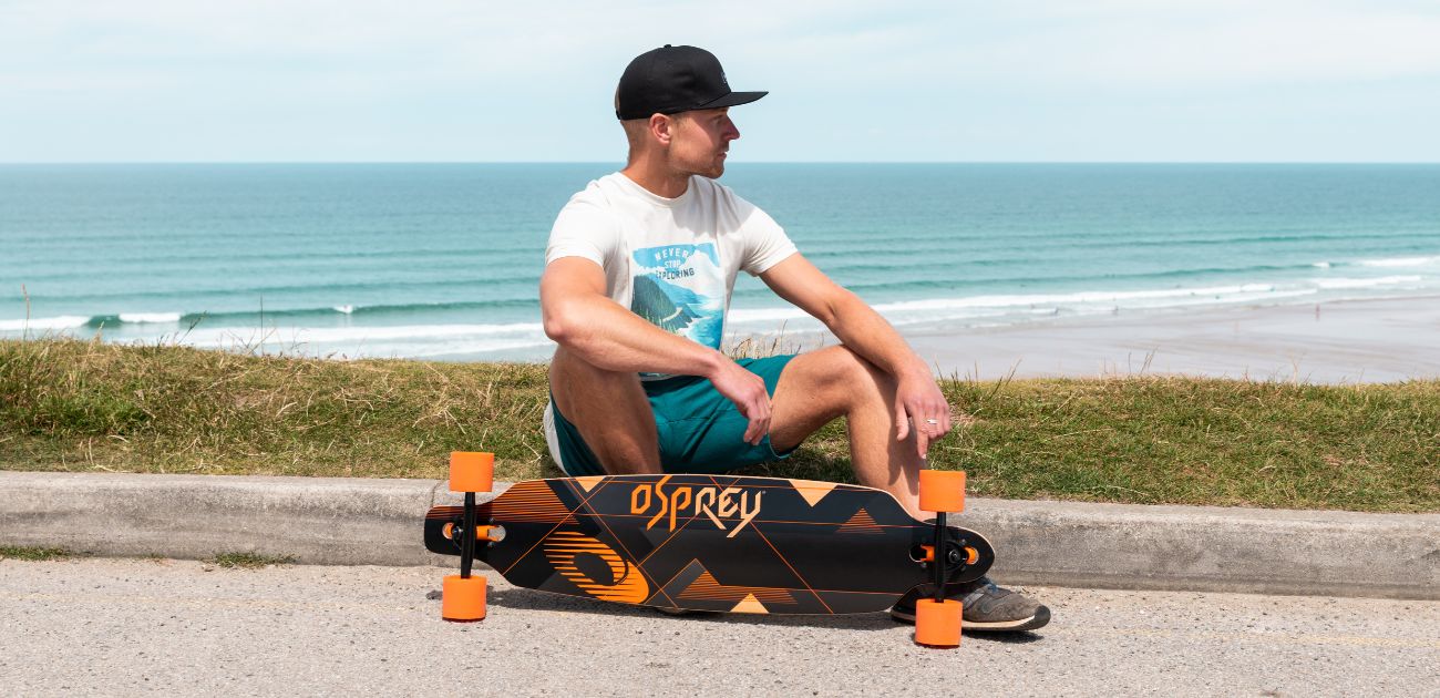 a man sat on the floor with an Osprey skateboard. The sea is behind himm