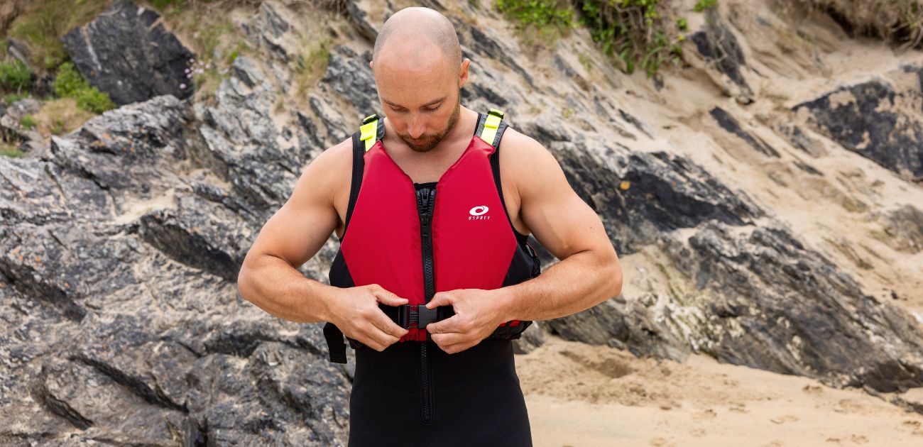 a man standing in front of some rocks fastening a red Osprey PFD