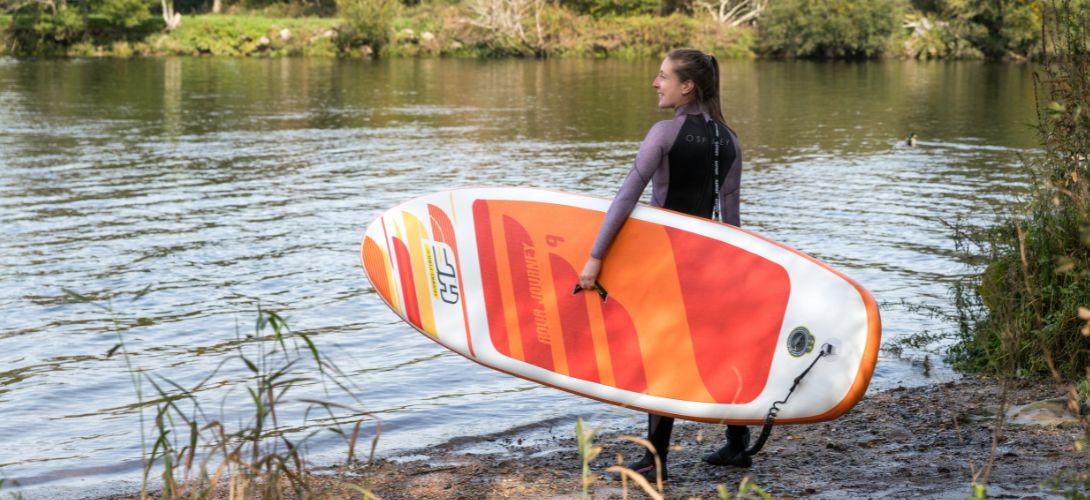 A photo of a woman carrying an orange Aqua Journey paddleboard towards a river