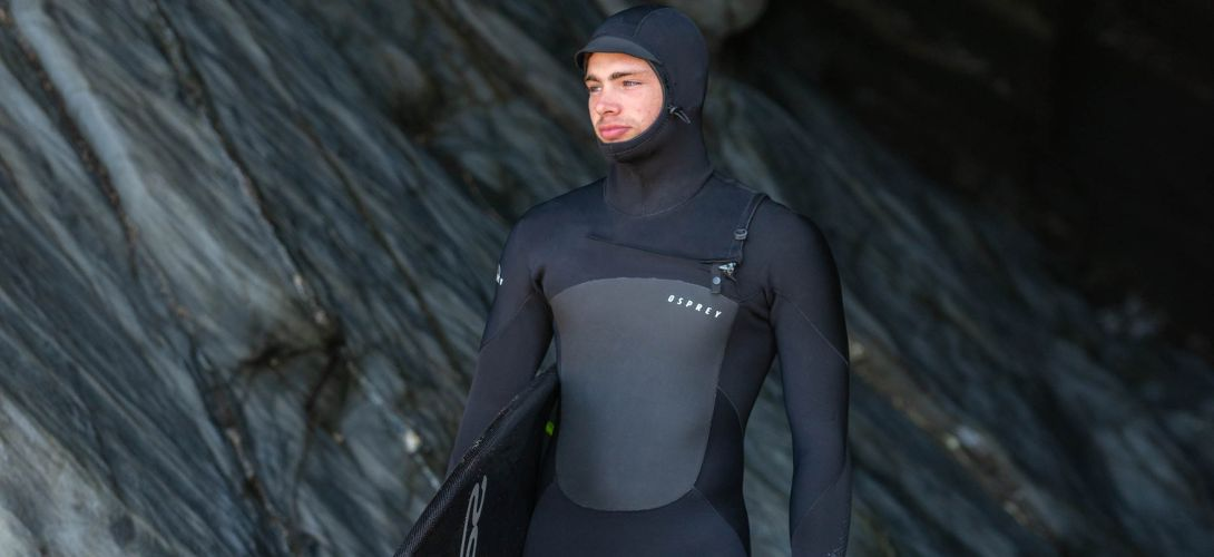 A photo of a man wearing a black hooded wetsuit stood in front of some dark grey rocks