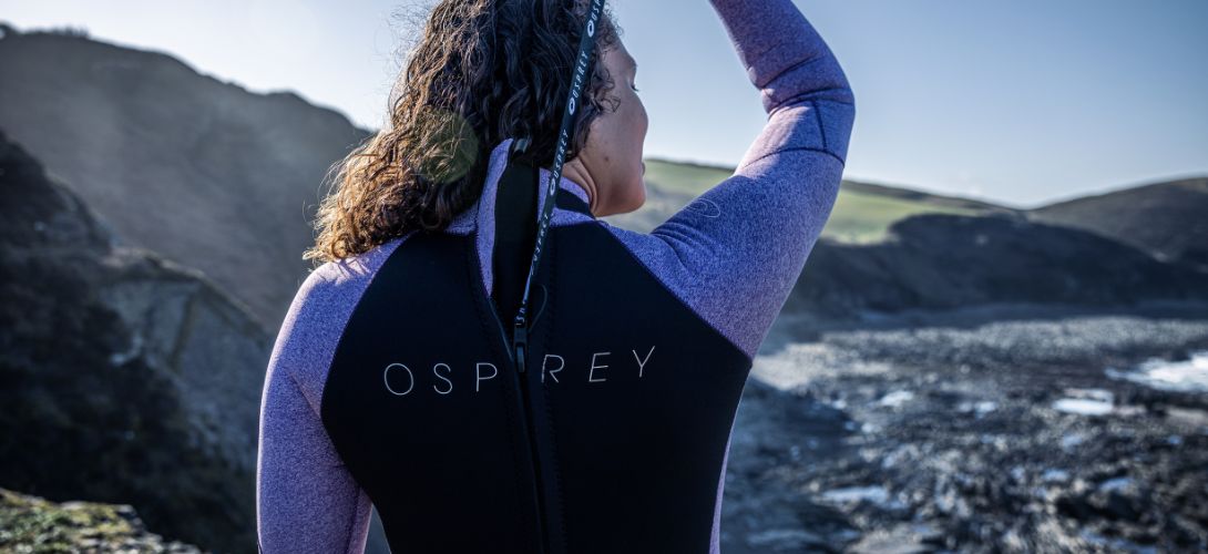 A photo of a women at sunrise pulling the back zip up on an Osprey purple and black wetsuit