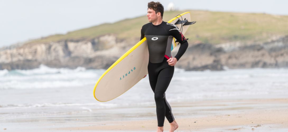 A photo of man running across a beach carrying a yellow surfboard and wearing a red and grey Osprey wetsuit