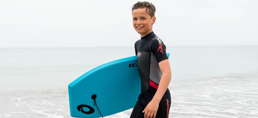 A photo of a boy in front of the sea. He is wearing a red and grey Osprey wetsuit and carrying a blue bodyboard