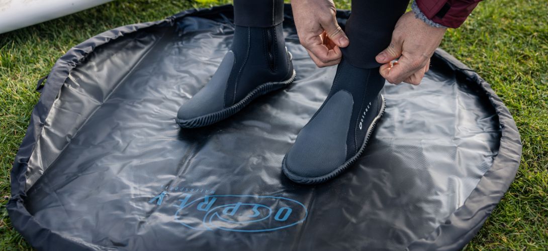 A photo of someone fastening black wetsuit boots. They are standing on a black changing mat