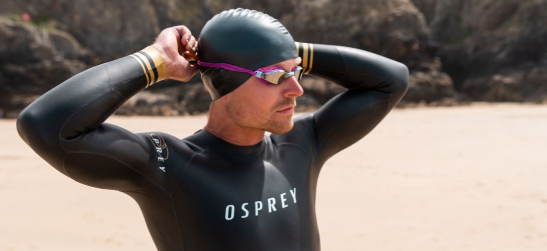 A photo of a man stood on a beach. He is wearing a triathlon wetsuit and swimming hat and adjusting a pair of pink goggles