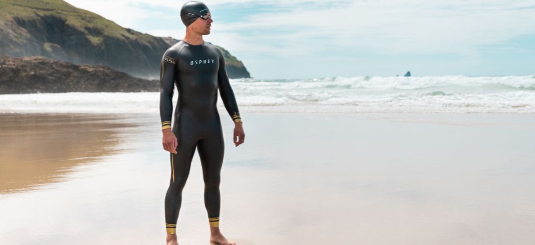 A photo of a man standing on a beach in a trisuit, swimming hat and goggles.