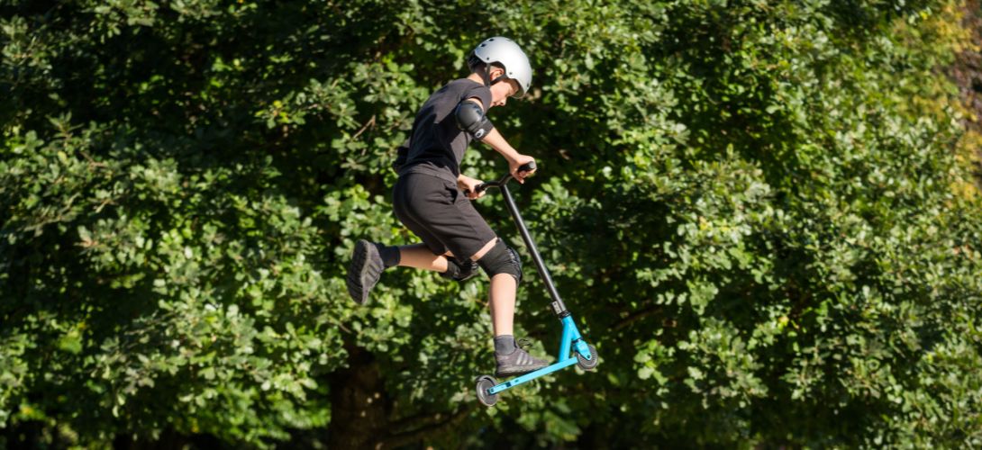 A photo of a young boy performing a trick on a blue stunt scooter. He is in the air and the background is trees