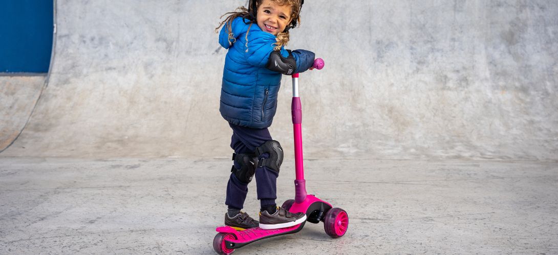 A photo of a young boy standing in a skate park with a pink three wheeled scooter