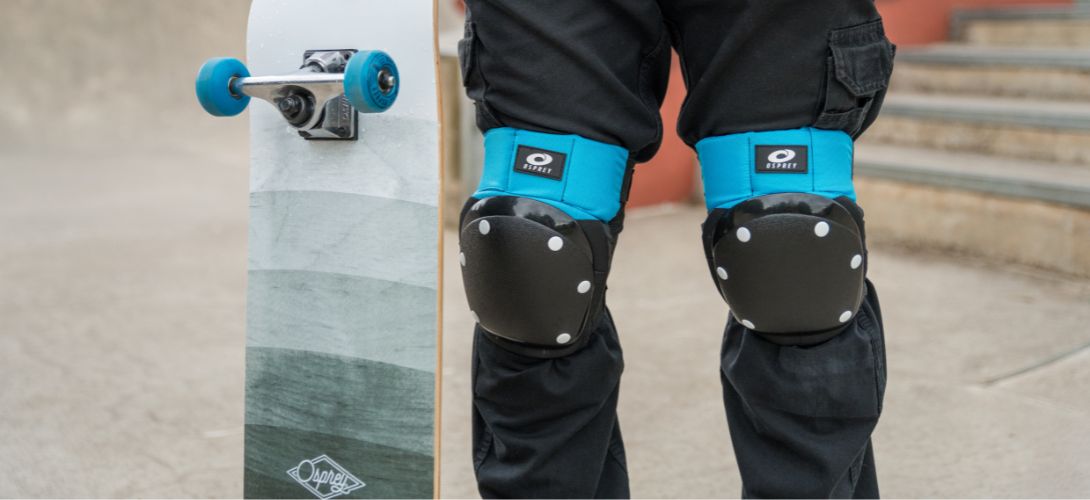 A close up photo of someone wearing an Osprey knee pad set, he is holding a grey Osprey skateboard at his side