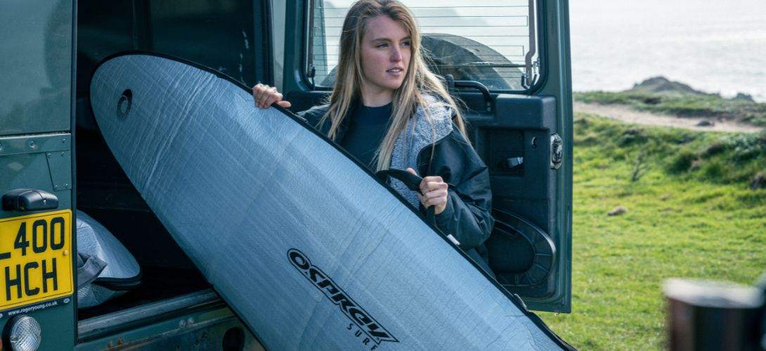 A photo of a blonde woman taking her surfboard bag out of the black of a grey Land Rover