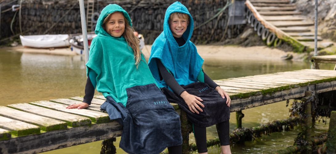 A photo of a girl and a boy sat on a small jetty wearing Osprey towel ponchos. They are both smiling and sat with their hoods up