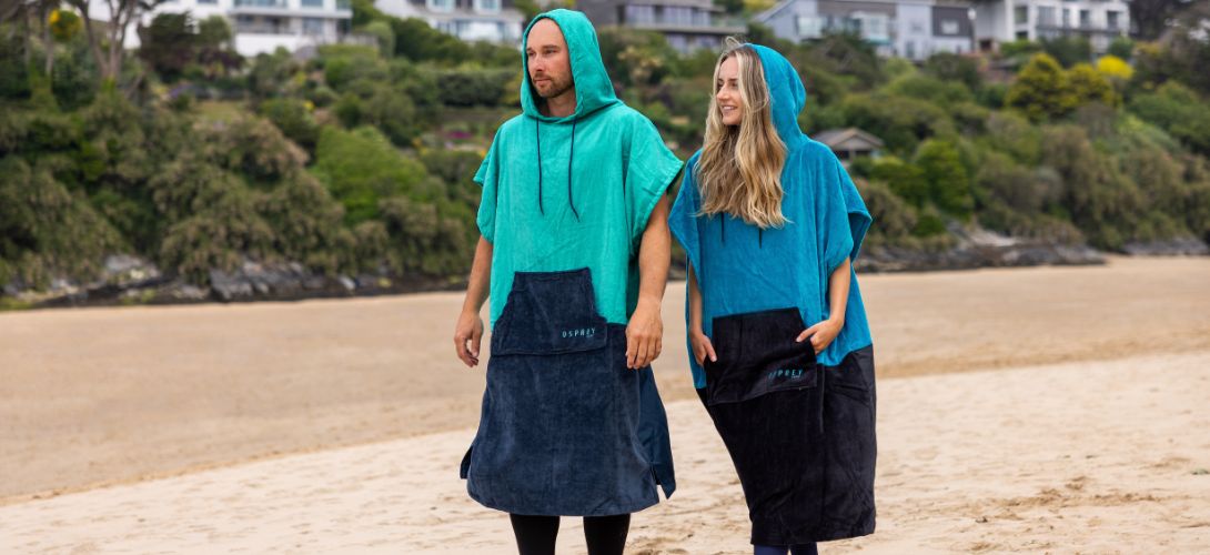 A photo of a man and woman walking along a beach wearing towel ponchos with their hoods up