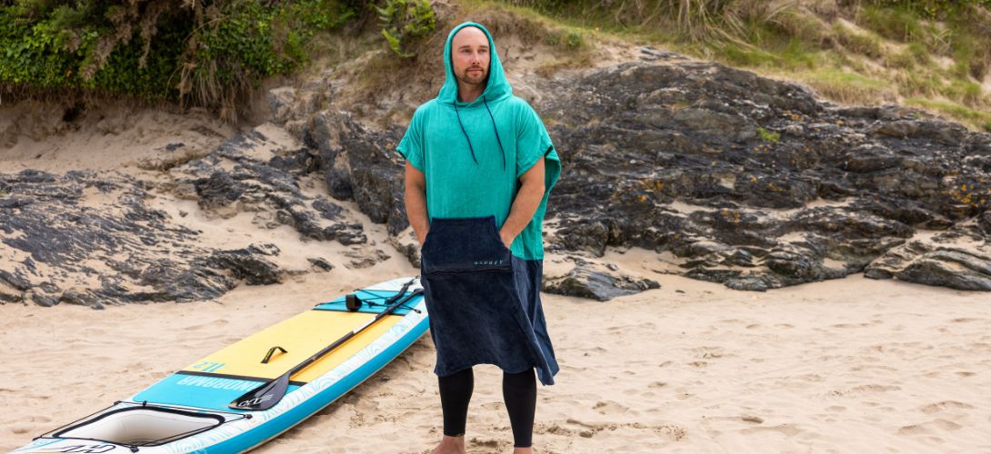 A photo of a man stood on a beach in a teal changing poncho. He has a paddleboard laid next to him on the floor