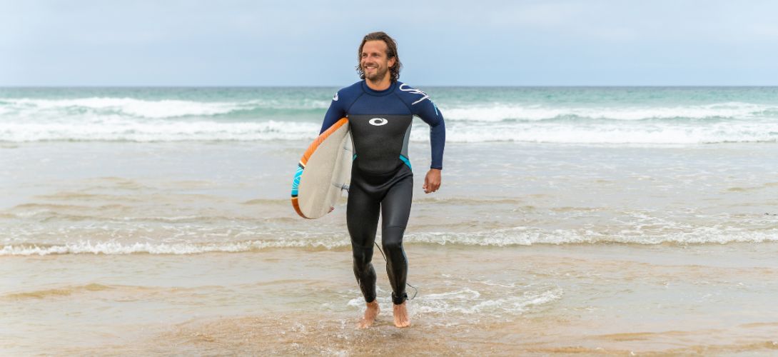 A photo of a man running out of the sea. He is wearing a navy wetsuit and carrying a foam surfboard