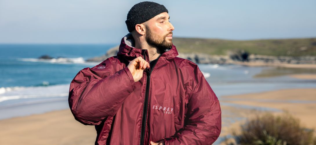 A photo of a man stood in front of a beach. He is wearing a black beanie hat and zipping up a maroon changing robe