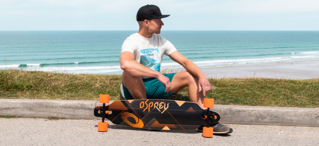 A man sitting on a kerb in front of the sea, he has an orange and black Osprey longboard in front of him