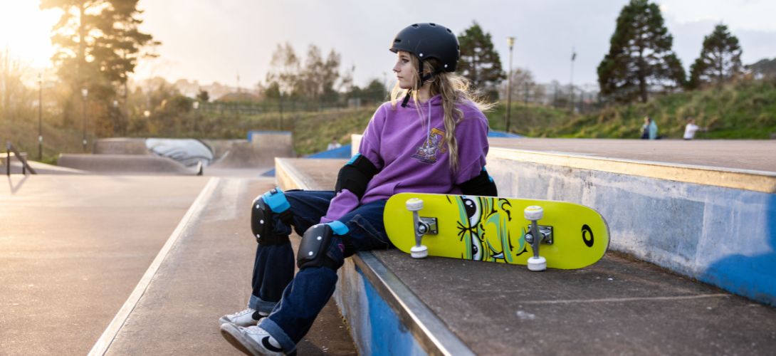 A photo of a girl sat at a skatepark at sunset, she has a yellow doublekick skateboard rested next to her