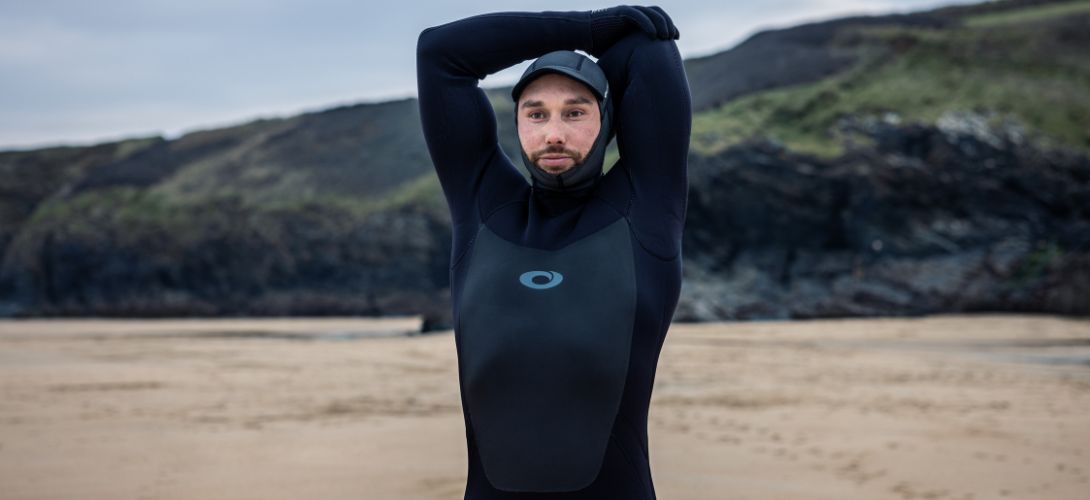 A photo of a man stood on the beach stretching and wearing a black Osprey wetsuit with a hood