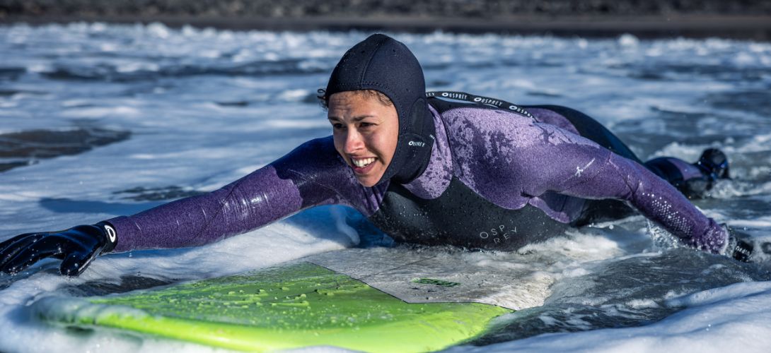 A photo of a female surfer paddling in the water wearing an Osprey wetsuit and hood.